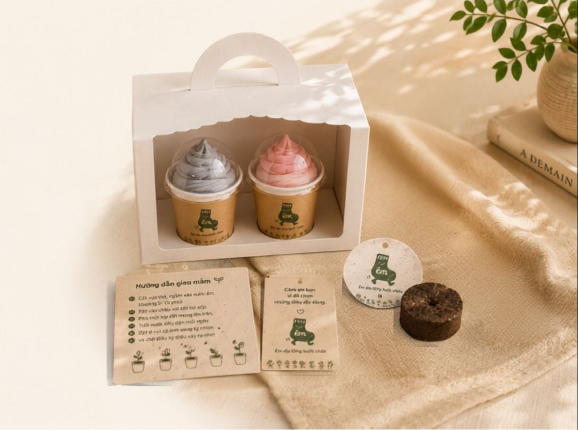 Hình ảnh Section Packaging Story giới thiệu hộp quà trải nghiệm của thương hiệu êm gồm 2 đôi tất cuộn hình bánh cupcake đặt trong hộp giấy quai xách màu trắng kèm giấy hướng dẫn, thẻ gieo mầm seed paper và viên đất nén mini trên nền vải thô